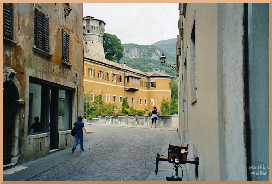 Ockerfarbenes Schloss mit Festungsturm darüber aus Pflasterasse vor Brücke betrachtet, Rovereto
