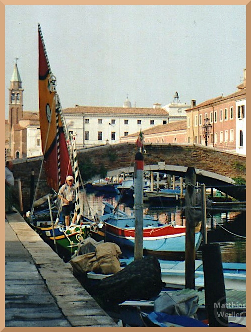 Barke mit buntem Segel vor Kanalbrücke und spitzem Kirchturm in Chióggia