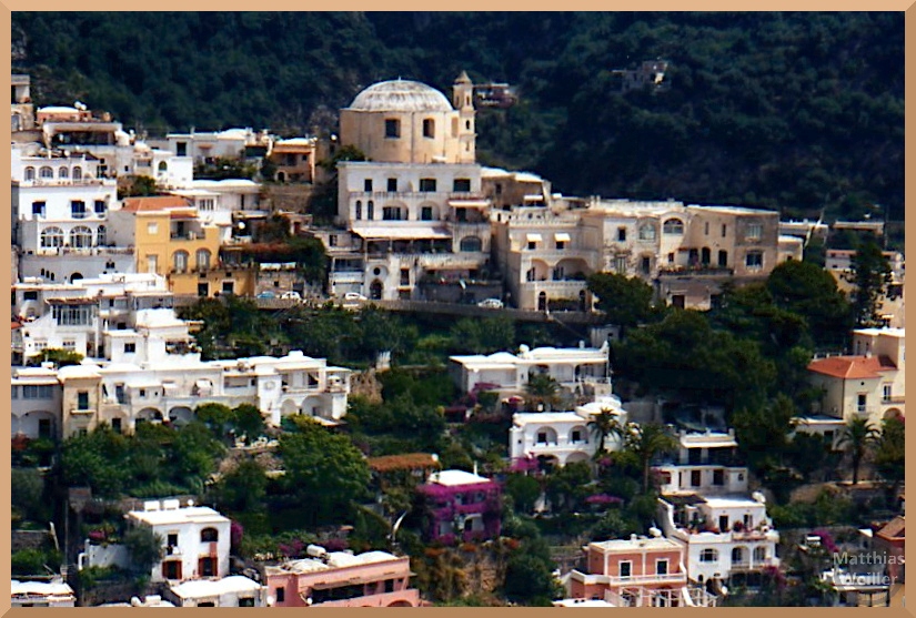 Frontansicht von Positano mit Kuppelbau, grün/weinroter Bewuchs zwischen und an Häusern, weiße Fassaden