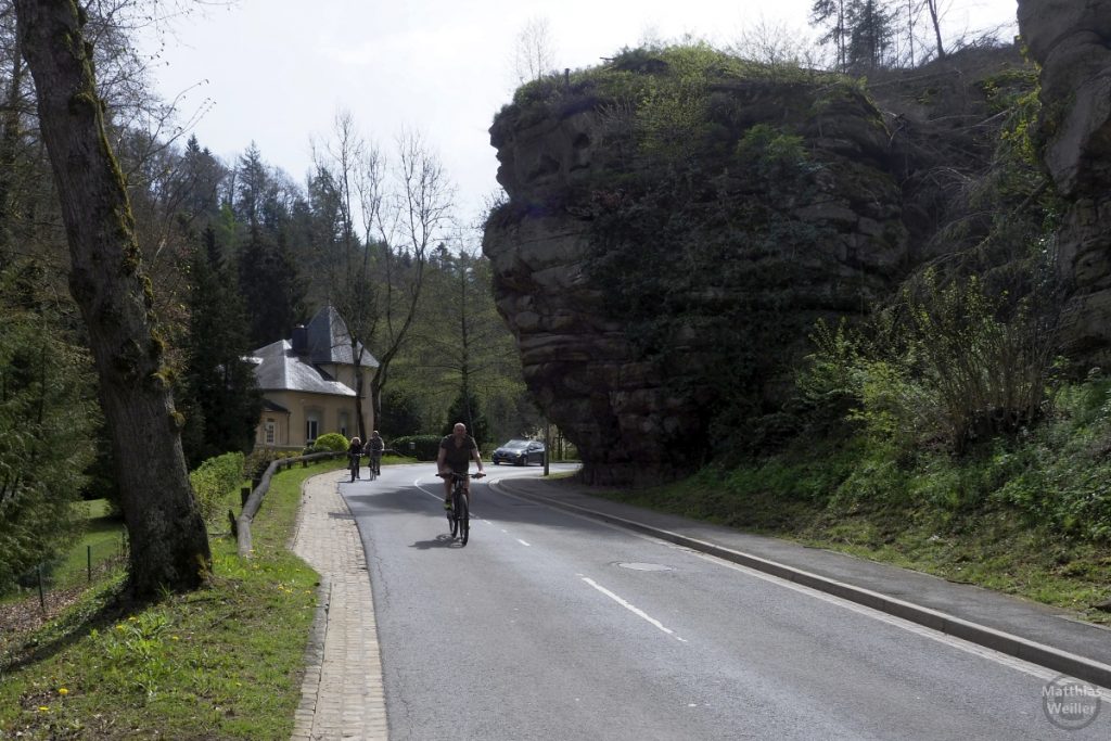 Straße mit Radfahrern und überhängendem Fels an Kurve, bei Larochette