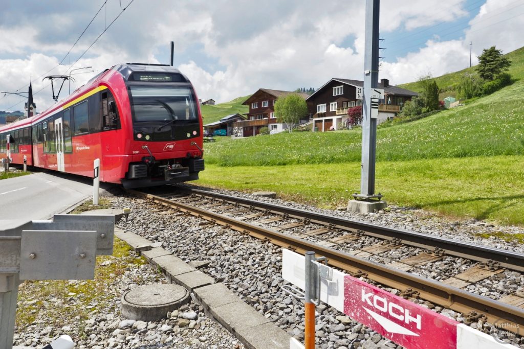 Schweizer Bähnli an Bahnübergang mit typischen Landhäusern und grünem Berghang