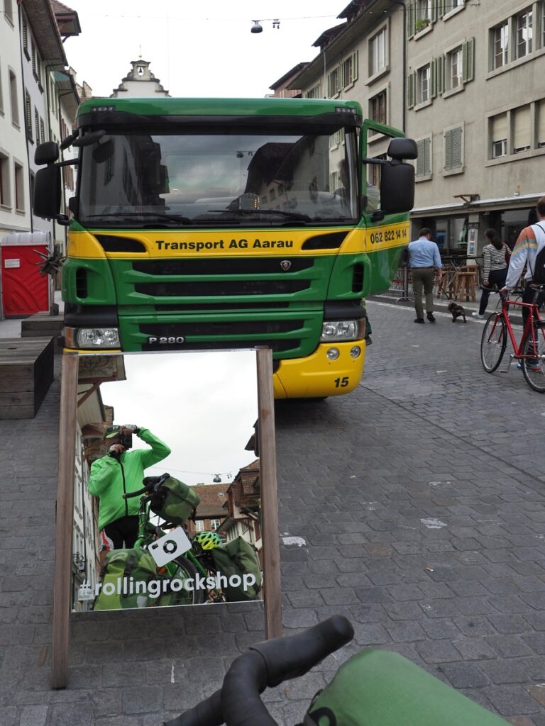 Fotograf mit grüner Jacke, grünem Reisevelo vor Spiegel mit grün-gelbem Transporter im Hintergrund