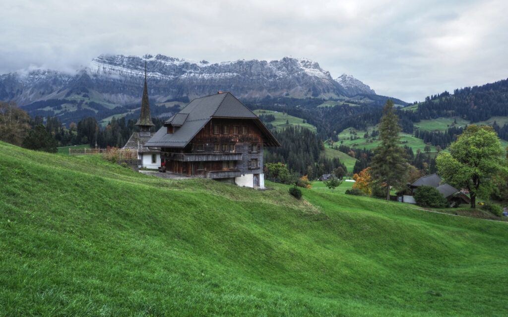 Schrattenfluh mit Schneegriesel vor Bauernhaus mit Kapelle und spitzem Turm, grüne Bergwiese, düster