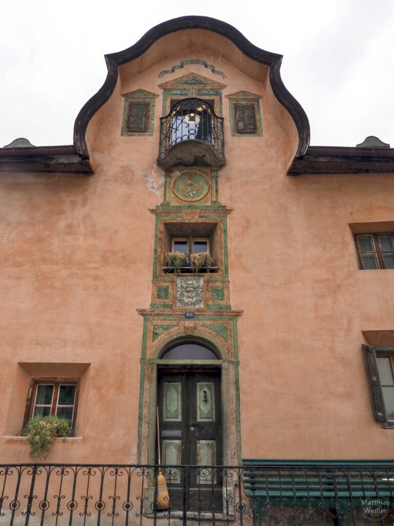 Engadinerhaus, ocker/grün geschwungene Dachform, gestaffelte Tür/Fenster/Balkon, Ardez