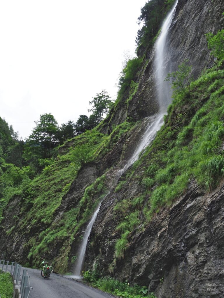 Wasserfallstrahl über Fels an Piste mit Velo