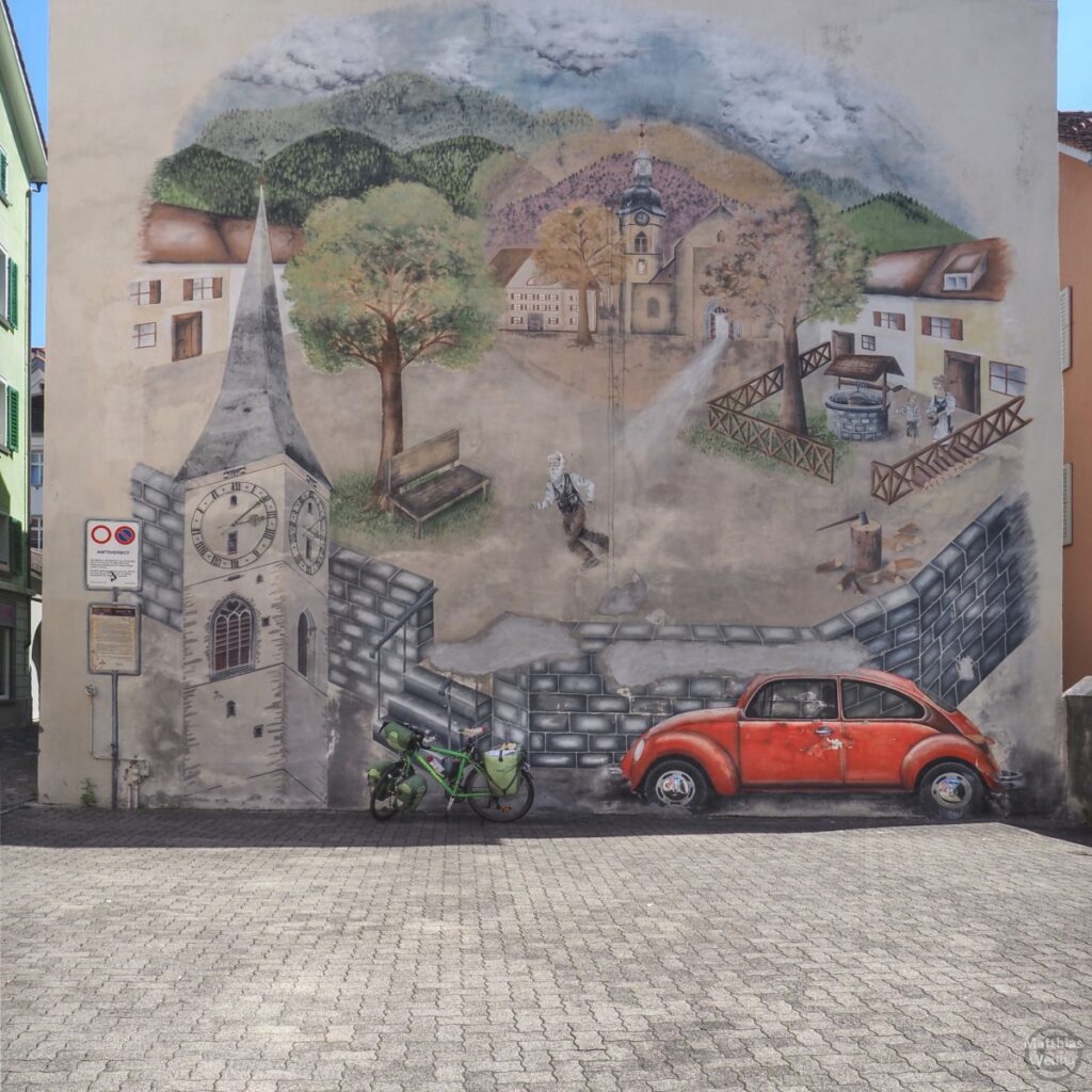 Fassadenbild mit Geschichtshintergrund der Stadt Chur, gemalter VW-Käder rot, grünes Reisevelo real
