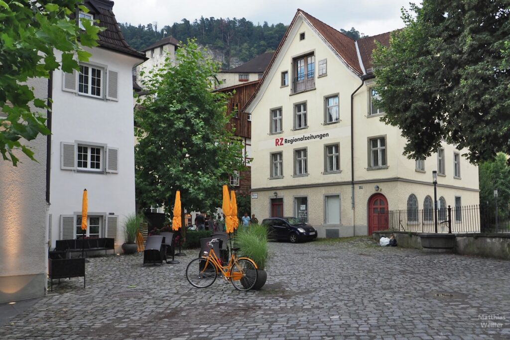 Bild von Platz mit orangenem Velo, Sonnenschirmen und Haus RZ Regionalzeitung