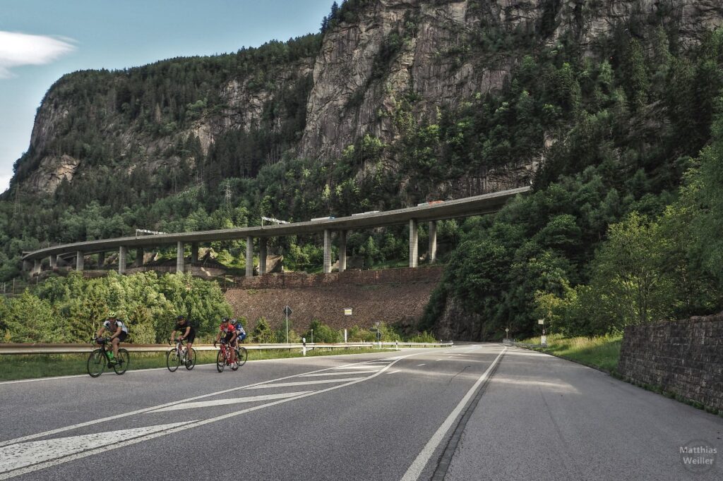 Valle Leventina mit Straße, vier Rennradlern und Autobahnbrückentrasse vor Felshang