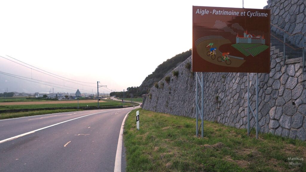 Straße, Mauer und Schild "Aigle - Patrimoine et Cyclisme"