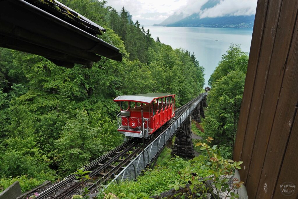 Rote Standseilbahn Giessbach von 1879 auf Schienenviadukt durch grünen Wald mit See im Hintergrund