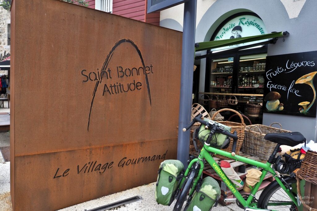 Grünes Reiserad vor Regionalproduktladen und Rostmetallfläche "Saint Bonnet Attitude - Le Village Gourmand"