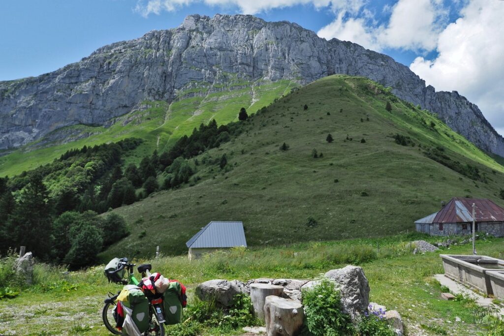 gebogene Felsfront über grünem Bergfundament, mit Reiserad, Brunnen, Bauernhütten, Col de Chérel