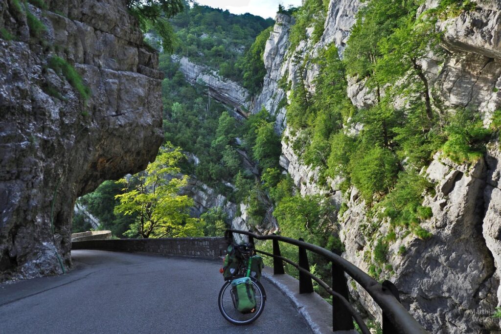 Schlucht mit geschichten Felsen und Überhängen, von grünen Bäumen durchsetzt, mit Reiserad auf Straße