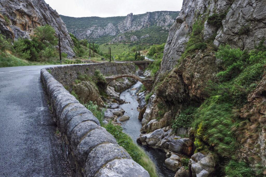 schluchtiges Tal mit verkarsteten Bergen, Straße mit Randmauer, Fluss Agly, und alter Bogenbrücke