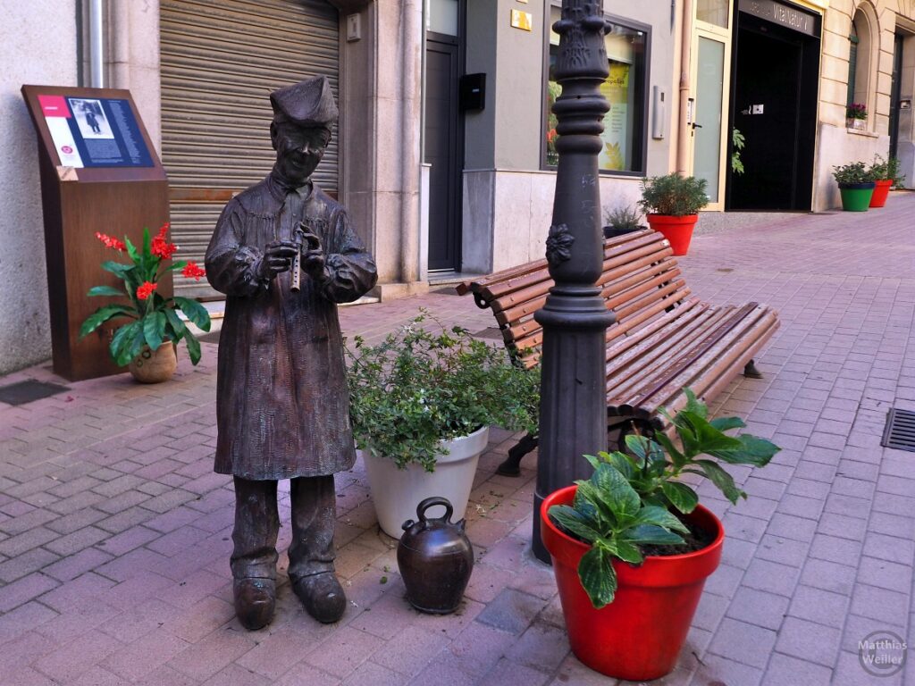 Skulptur eines legendhaften Flötenspielers in Sant Hilari Sacalm, vor Sitzbank und mit Blumentöpfen