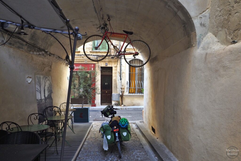 Rennrad mit Weinflache unter einem Hausbogen hängend, grünes Reiserad darunter (Avignon)