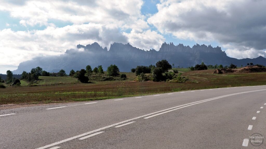 Gebirgskette der Muntanya de Montserrat, die aus einer Ebene emporsteigt, mit Wolken, Straße im Vordergrund