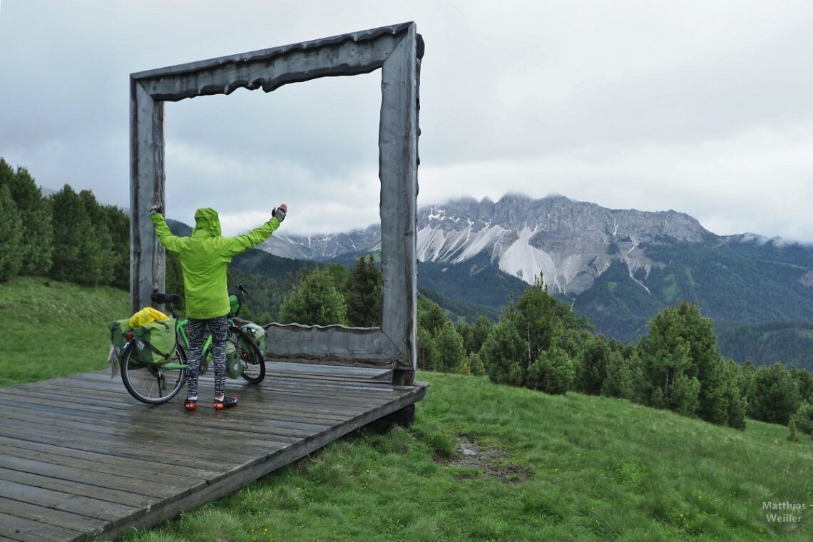 Rahmenblick auf Bergpanorma am Plose, teils in Wolken, mit Velo, Selbstporträt in Regenjacke von hinten