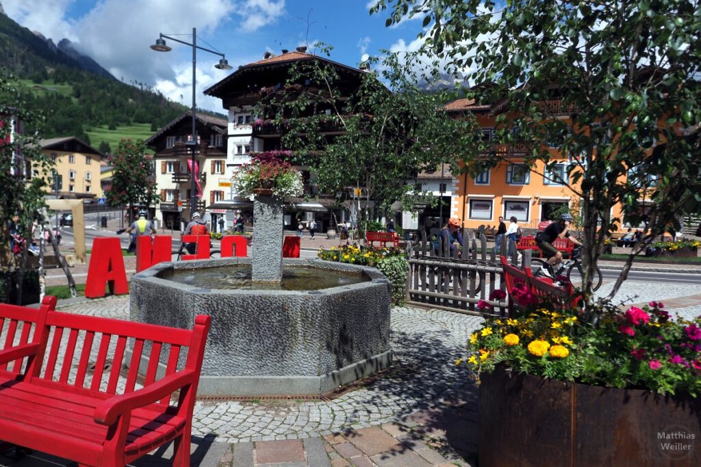 Moena, Ortsbild mit Brunnen, roter Bank, rotem Ortsnamen, Radler im Hintergrund