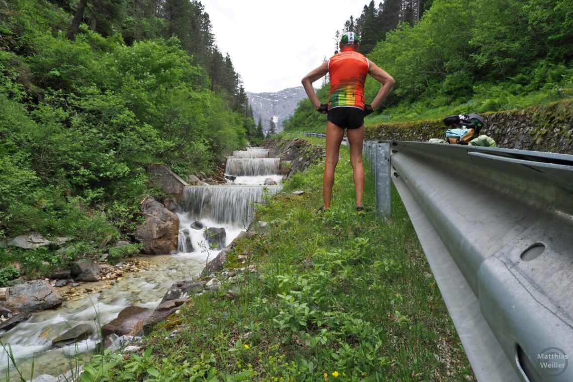 Selbstporträt von hinten neben Kaskadenbach, neben Leitplanke und Velo