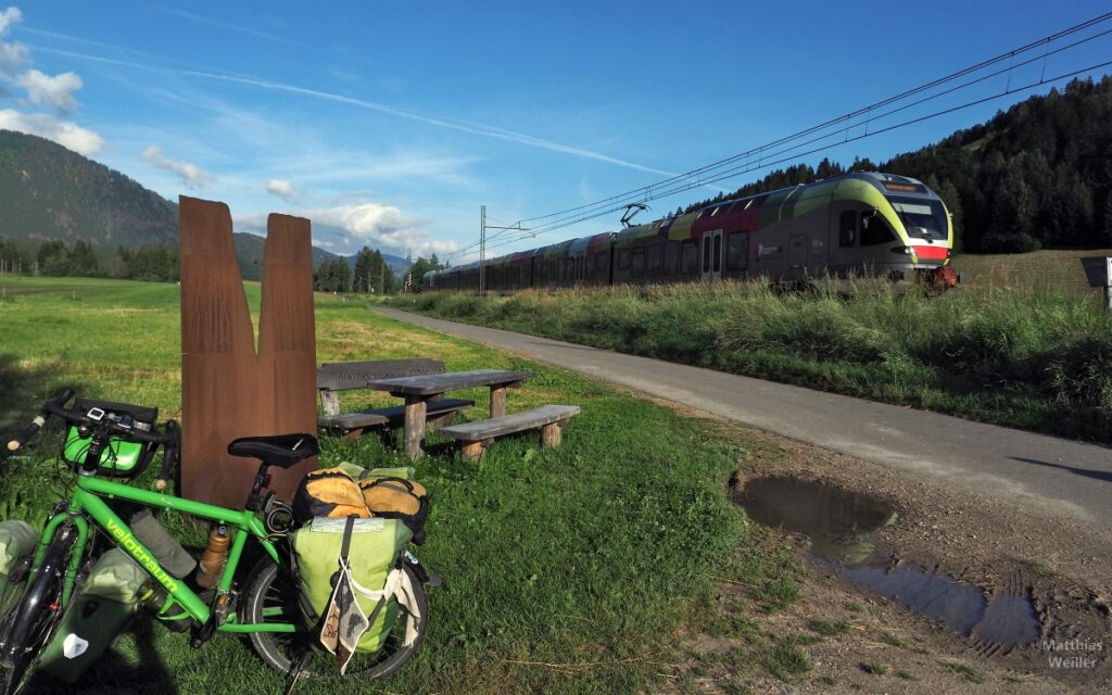 Skulptur Drauquelle mit Velo am Radweg, vorbeifahrender Zug