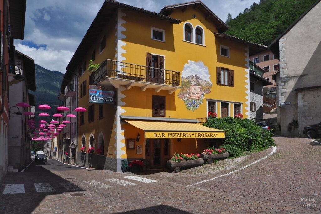 Ockerfarbens Haus mit Malerei, pinkfarbene Regenschirme über Ortstraße