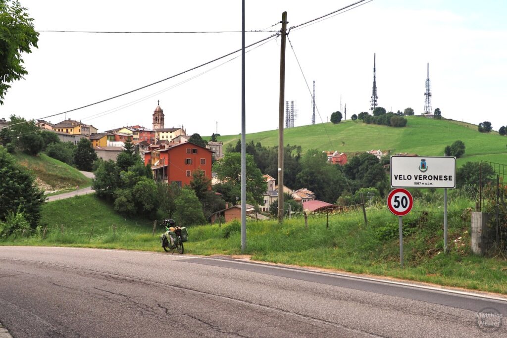 Ortsbild Velo Veronese mit Ortschild, Sendemasten auf Hügel und Velo