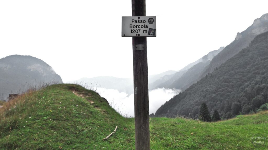 Kleines Schild "Passo Borcola 1207 m" vor Hügel-/Wolkenkulisse