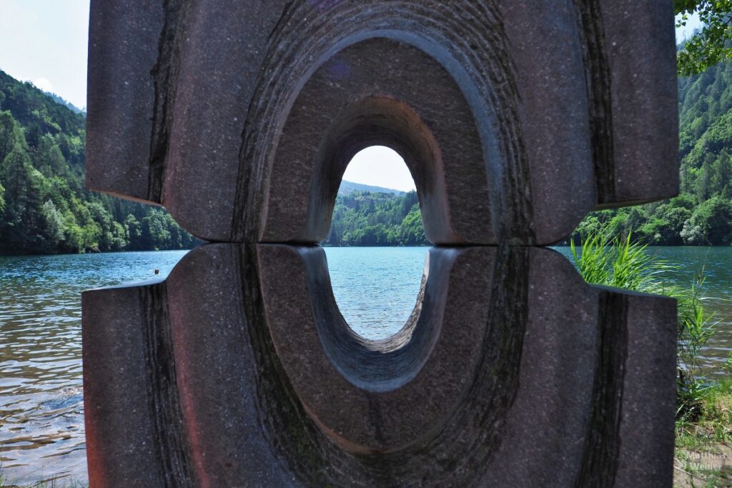 Blick durch Steinskulptur mit ovalen Loch auf Lago di Lases