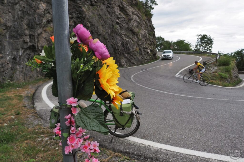 Spitzkehre mit Rennradler, Auto, Velo, Blumen an Pfahl